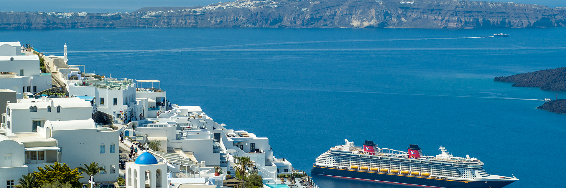White hillside buildings and blue-domed church overlooking the sea in Greece, with a cruise ship in the distance.