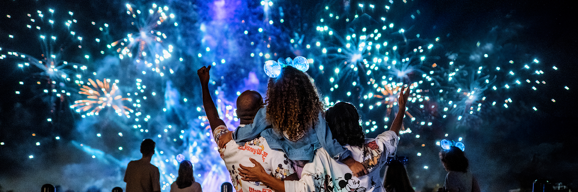 Guests watching Luminous, a nighttime fireworks display, over the water, with colorful bursts lighting up the sky.