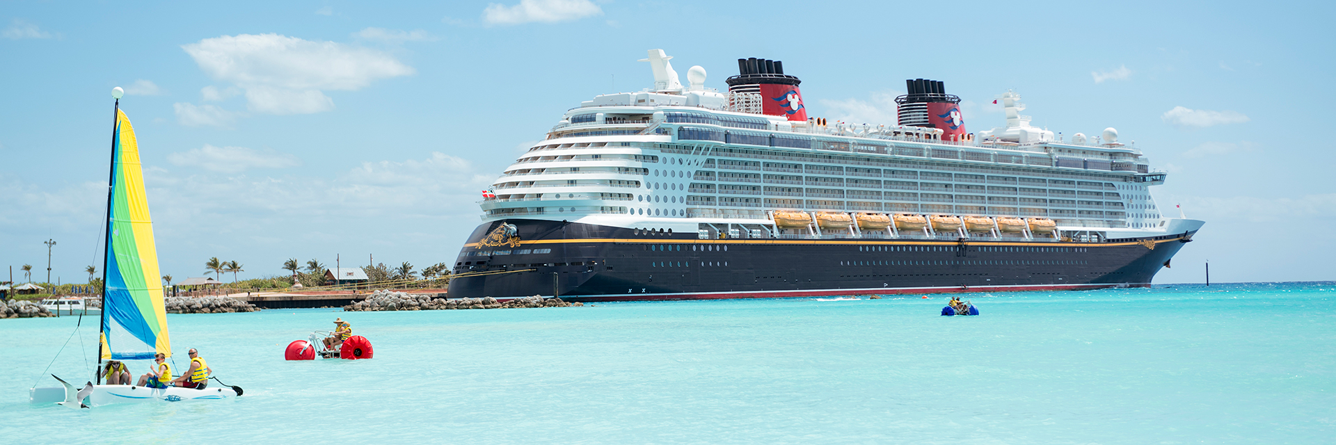 Cruise ship anchored near a tropical beach with turquoise water and small sailboats in the foreground.