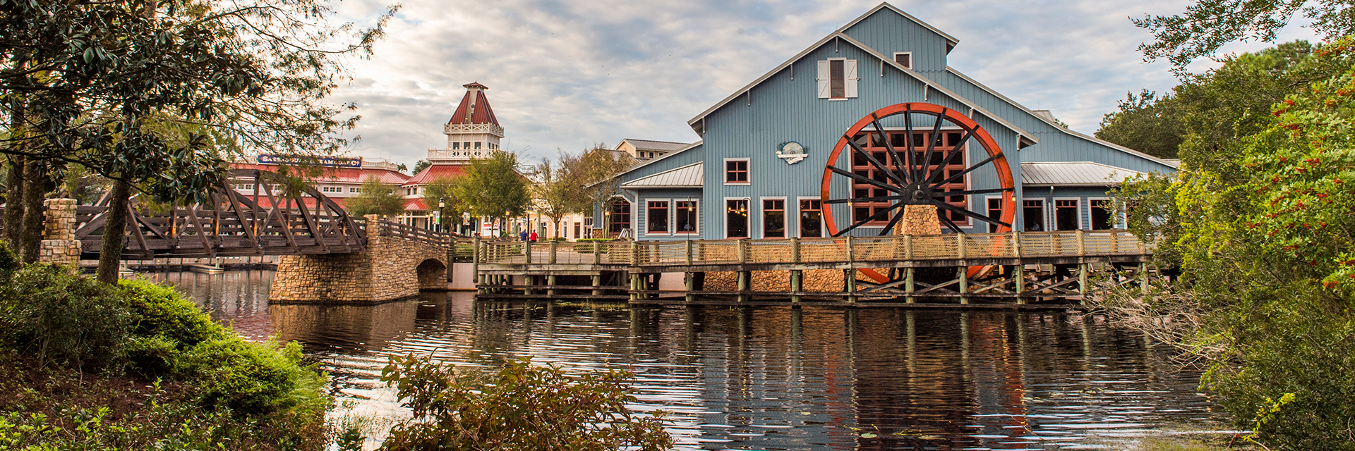 Blue waterfront building with a large water wheel beside a river and wooden bridge at Port Orleans Riverside.