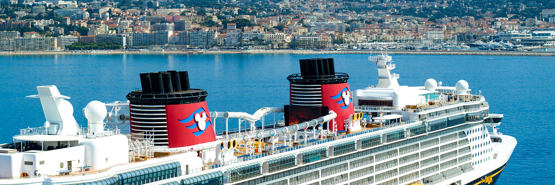 Large cruise ship sailing near a coastal French city, with yellow lifeboats and red funnels visible.