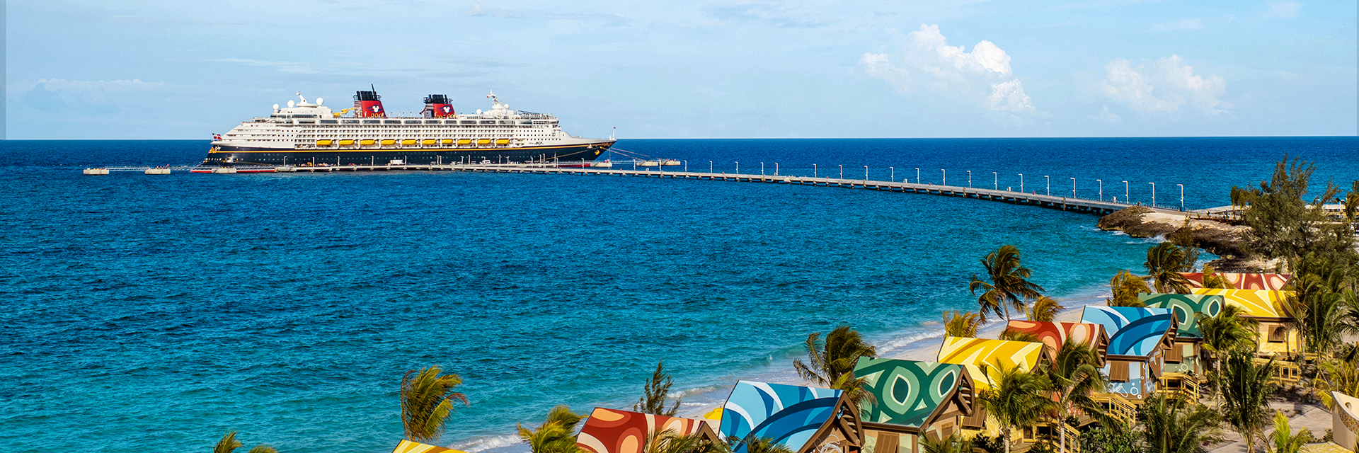 Cruise ship docked at a long pier offshore, with colorful seaside cabanas and palm trees along the beach.